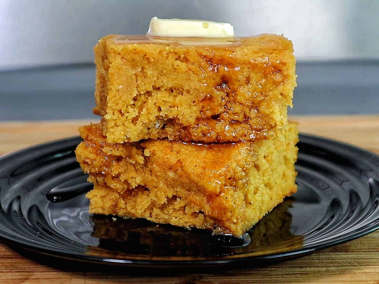 Two thick slices of brown sugar sweet potato cornbread stacked on a black plate, topped with melted butter and drizzled syrup. 