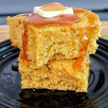 Two thick slices of brown sugar sweet potato cornbread stacked on a black plate, topped with melted butter and drizzled syrup.