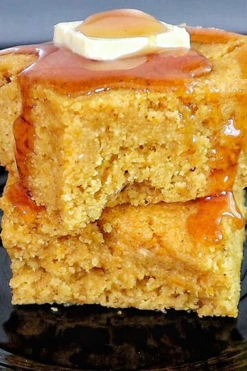 Two thick slices of brown sugar sweet potato cornbread stacked on a black plate, topped with melted butter and drizzled syrup.