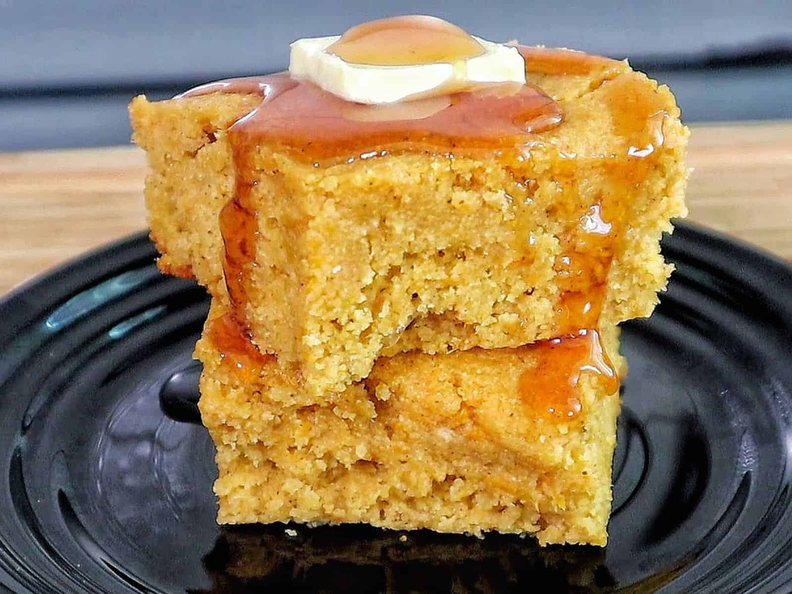 Two thick slices of brown sugar sweet potato cornbread stacked on a black plate, topped with melted butter and drizzled syrup. 