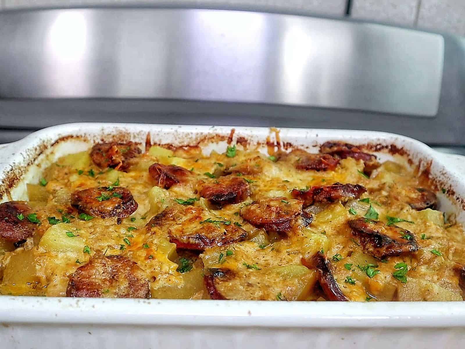 Cheesy sausage and potato casserole, featuring golden melted cheese, browned sausage slices, and tender potato cubes scooped from a baking dish.