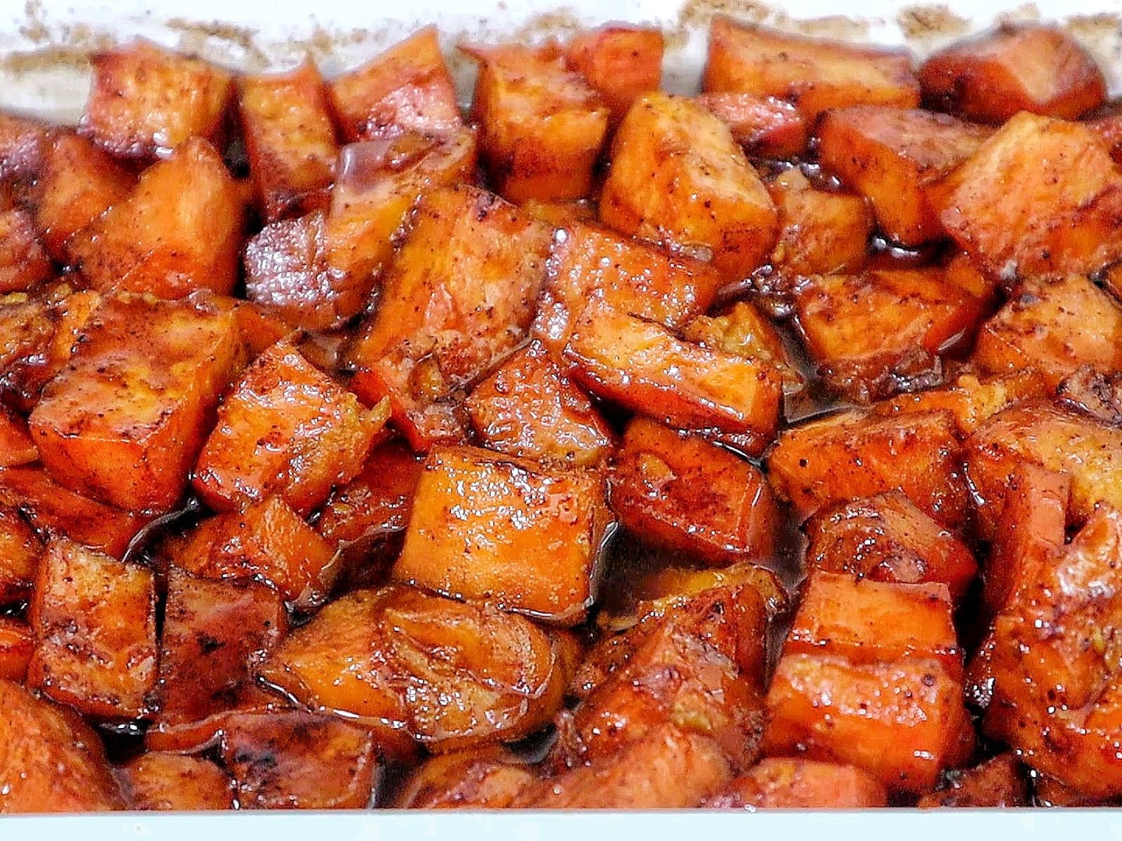 Southern candied yams featuring tender sweet potato chunks coated in a glossy brown sugar syrup with flecks of cinnamon and spices.