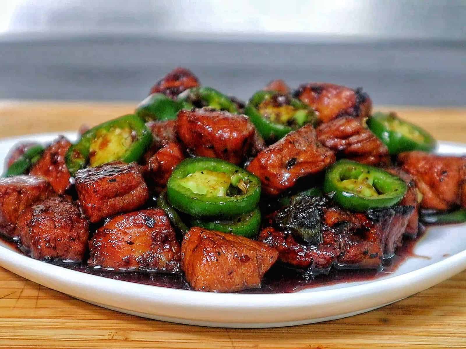 A plate of glazed pork cubes coated in a dark, sticky sweet sauce, topped with sliced fresh jalapeños.