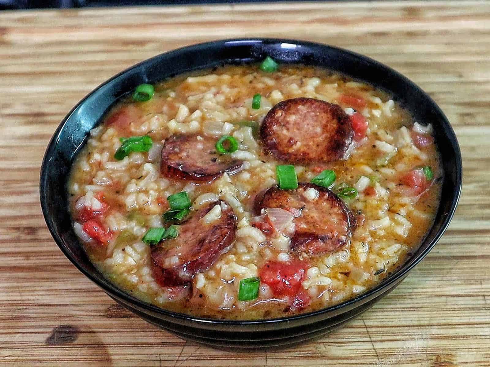 A bowl of jambalaya soup filled with tender rice, sliced smoked sausage, chunks of chicken, tomatoes, and vegetables in a rich, seasoned broth, garnished with chopped green onions.