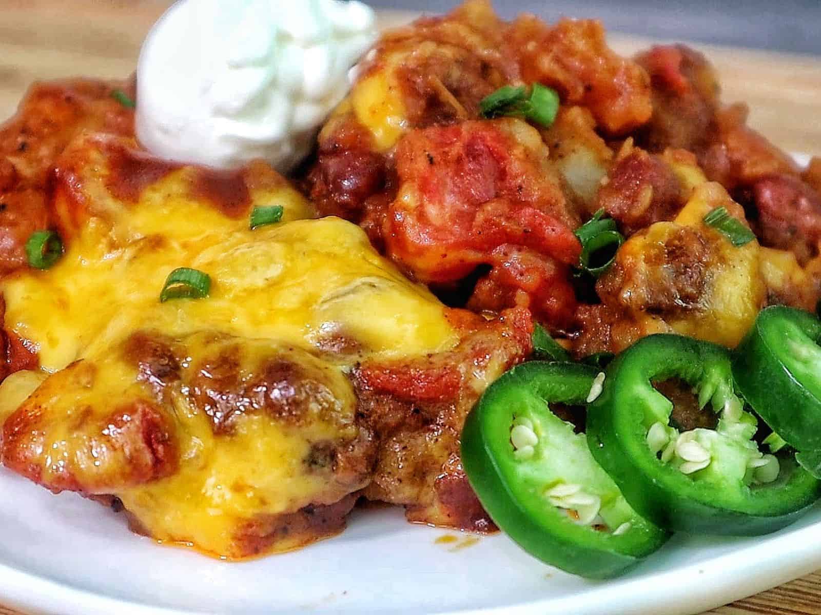 A plate of chili cheese potato bake topped with melted cheddar cheese, green onions, sliced jalapenos, and a dollop of sour cream, with tender potatoes coated in a chili tomato mixture.