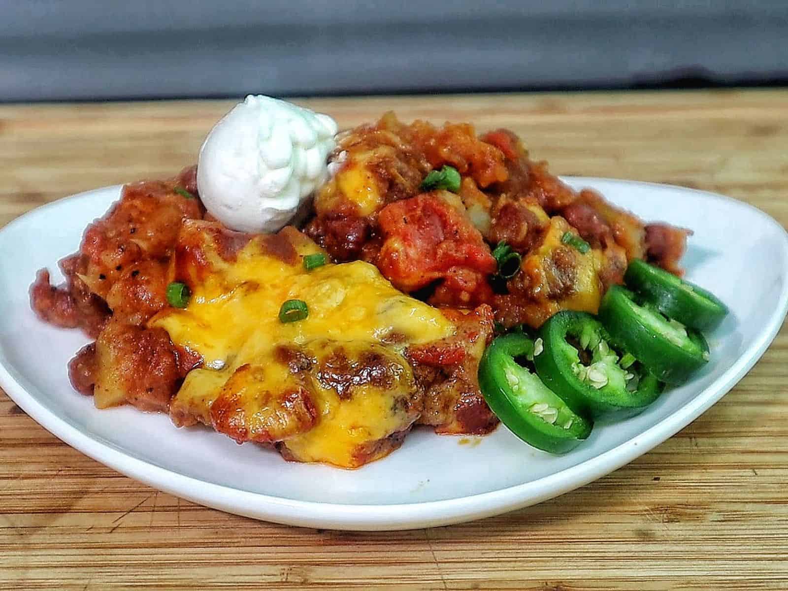 A plate of chili cheese potato bake topped with melted cheddar cheese, green onions, sliced jalapenos, and a dollop of sour cream, with tender potatoes coated in a chili tomato mixture.