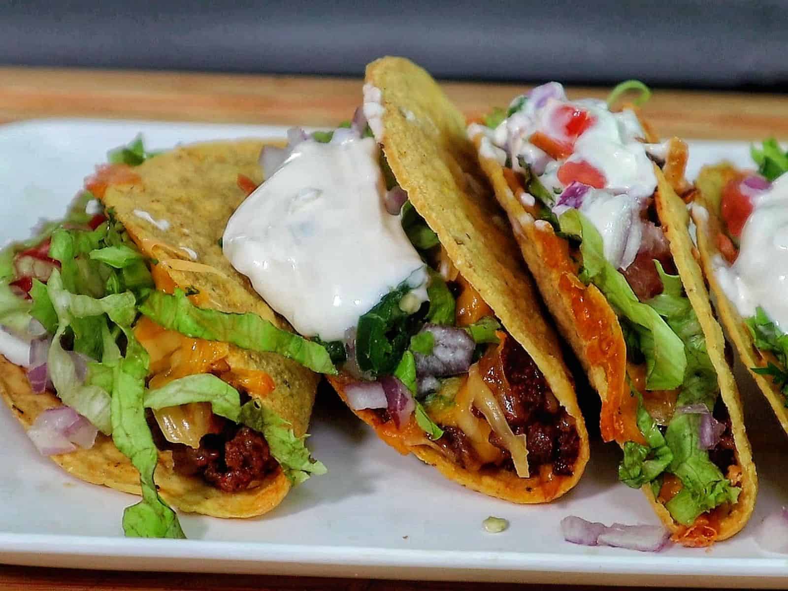 Cheesy oven baked tacos filled with seasoned ground beef, melted cheddar and smoked gouda cheese, topped with lettuce, tomatoes, onions, and sour cream in crispy taco shells.