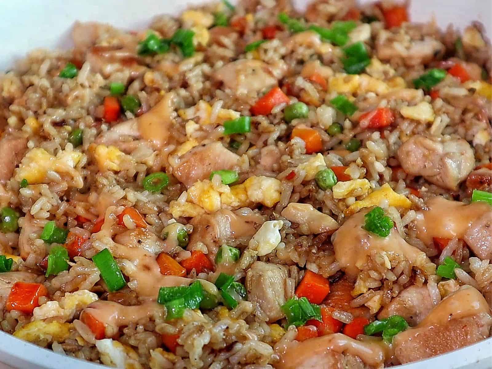 A plate of bang bang fried rice topped with green onions, featuring diced chicken, scrambled eggs, peas, carrots, and creamy sweet chili sauce drizzled throughout.