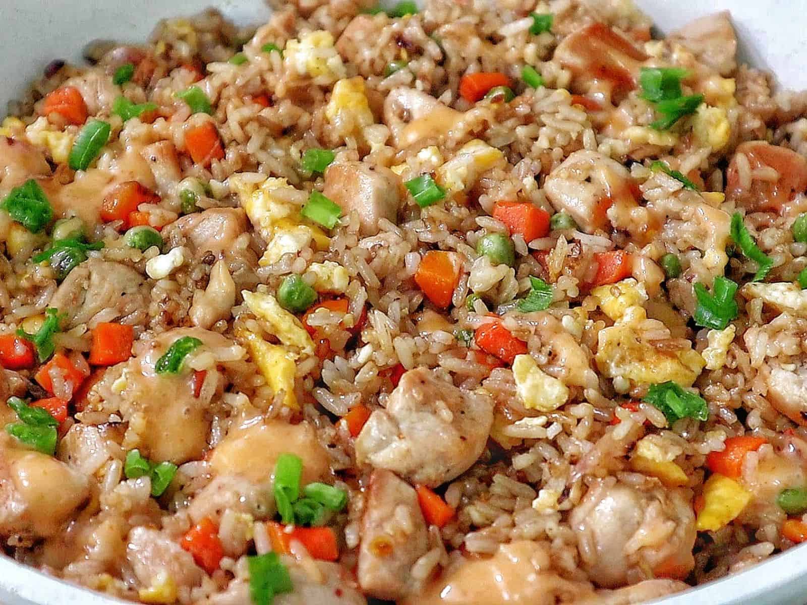 A plate of bang bang fried rice topped with green onions, featuring diced chicken, scrambled eggs, peas, carrots, and creamy sweet chili sauce drizzled throughout.