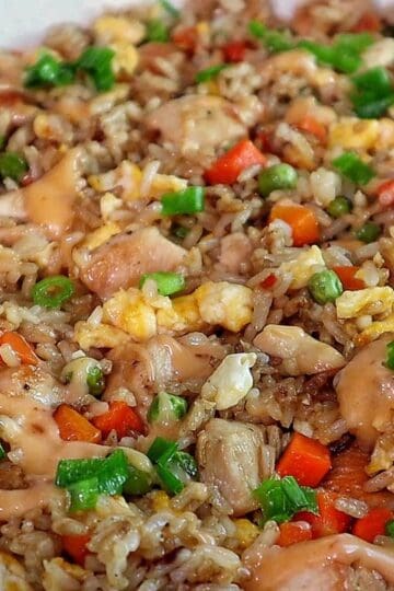 A plate of bang bang fried rice topped with green onions, featuring diced chicken, scrambled eggs, peas, carrots, and creamy sweet chili sauce drizzled throughout.