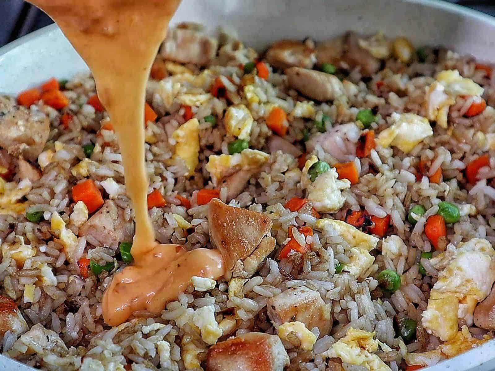 A plate of bang bang fried rice topped with green onions, featuring diced chicken, scrambled eggs, peas, carrots, and creamy sweet chili sauce drizzled throughout.