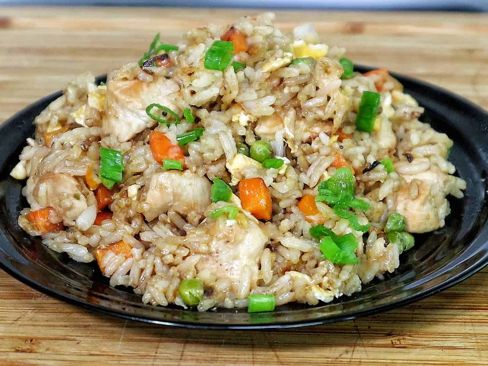 A plate of bang bang fried rice topped with green onions, featuring diced chicken, scrambled eggs, peas, carrots, and creamy sweet chili sauce drizzled throughout.