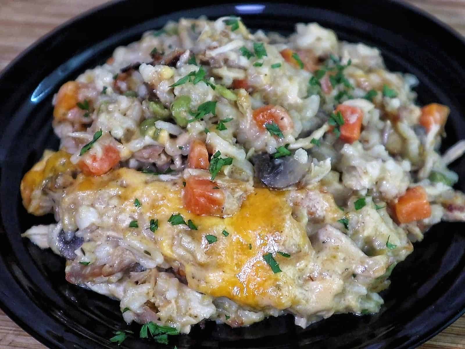 Creamy chicken and rice casserole topped with melted cheddar cheese, peas, carrots, and mushrooms with fresh parsley garnish.