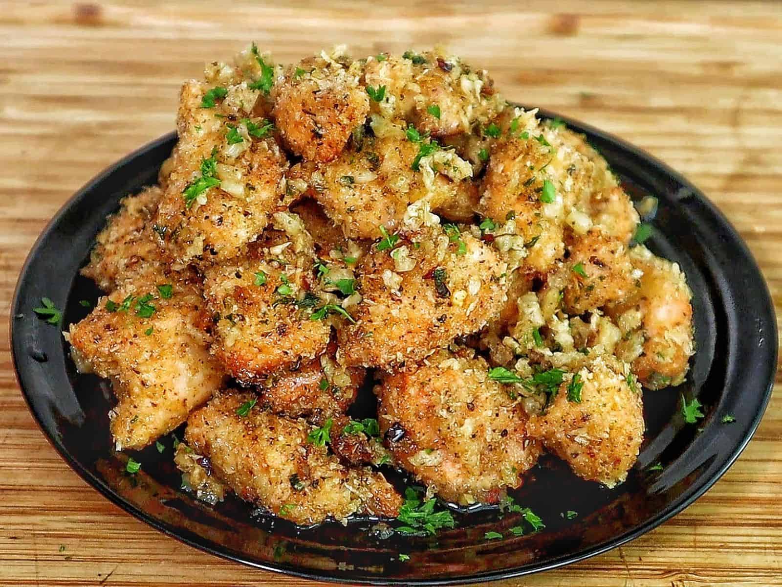 Golden brown garlic parmesan chicken bites piled on a black plate, coated in buttery garlic sauce and fresh parsley, with a crispy breadcrumb crust.