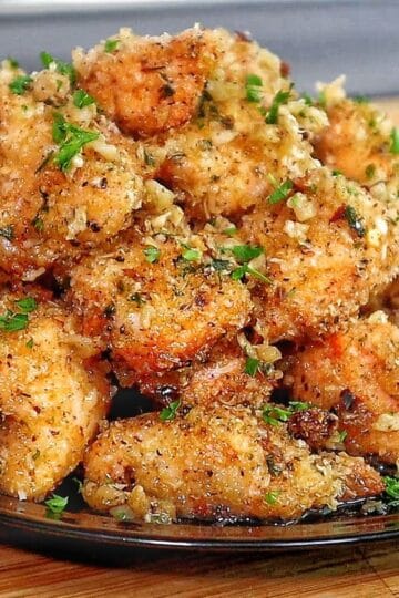 Golden brown garlic parmesan chicken bites piled on a black plate, coated in buttery garlic sauce and fresh parsley, with a crispy breadcrumb crust.