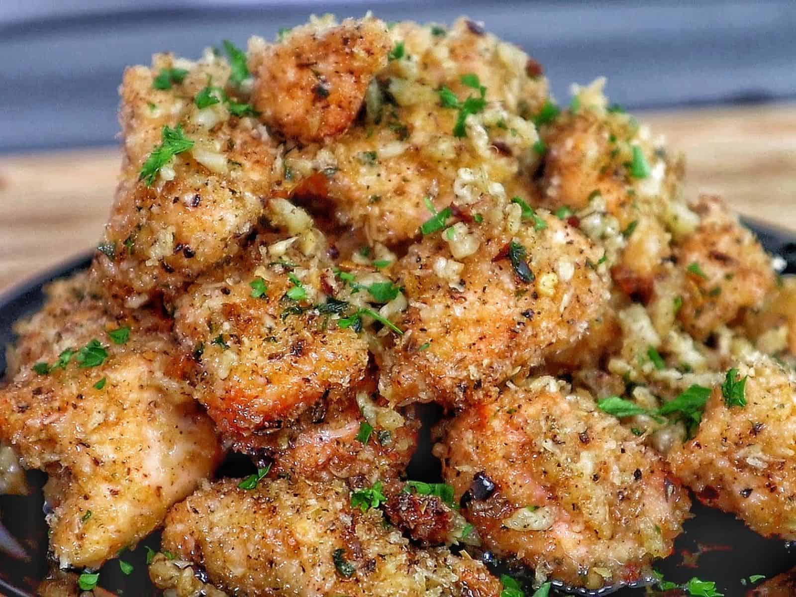 Golden brown garlic parmesan chicken bites piled on a black plate, coated in buttery garlic sauce and fresh parsley, with a crispy breadcrumb crust.