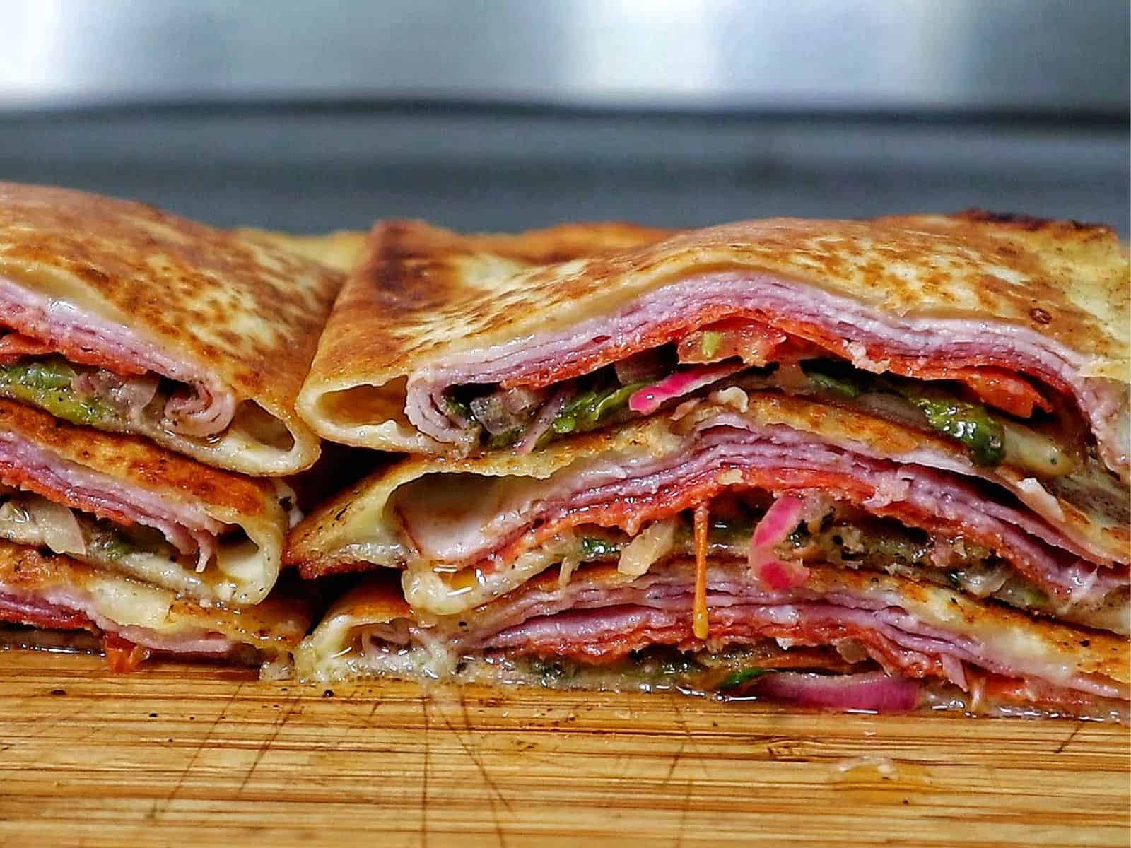 Toasted Italian sub wrap sliced in half, filled with layers of ham, salami, pepperoni, melted provolone cheese, lettuce, tomatoes, onions, and banana peppers.