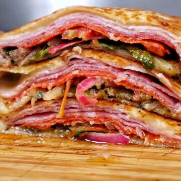 Toasted Italian sub wrap sliced in half, filled with layers of ham, salami, pepperoni, melted provolone cheese, lettuce, tomatoes, onions, and banana peppers.