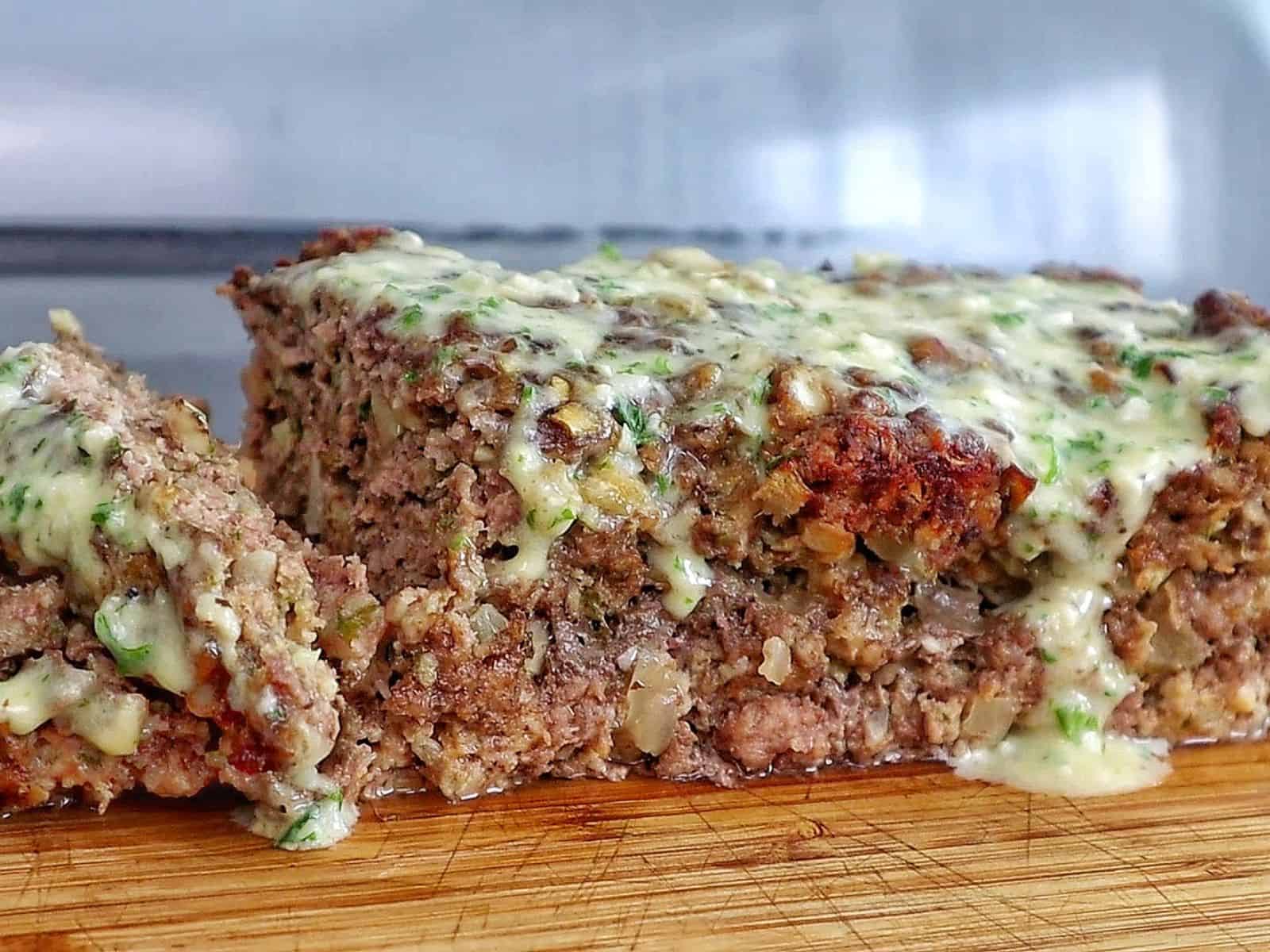 A thick slice of garlic parmesan meatloaf topped with a creamy garlic parmesan sauce and fresh parsley, served on a wooden cutting board with a juicy, tender center visible in every bite.