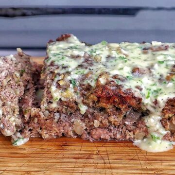 A thick slice of garlic parmesan meatloaf topped with a creamy garlic parmesan sauce and fresh parsley, served on a wooden cutting board with a juicy, tender center visible in every bite.