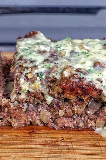 A thick slice of garlic parmesan meatloaf topped with a creamy garlic parmesan sauce and fresh parsley, served on a wooden cutting board with a juicy, tender center visible in every bite.