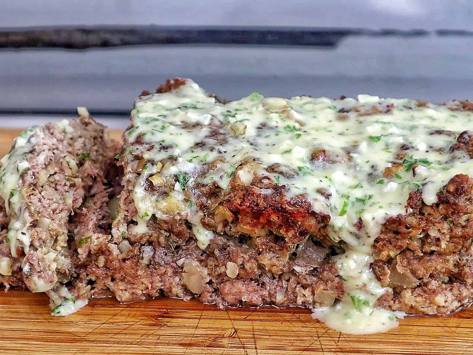 A thick slice of garlic parmesan meatloaf topped with a creamy garlic parmesan sauce and fresh parsley, served on a wooden cutting board with a juicy, tender center visible in every bite.