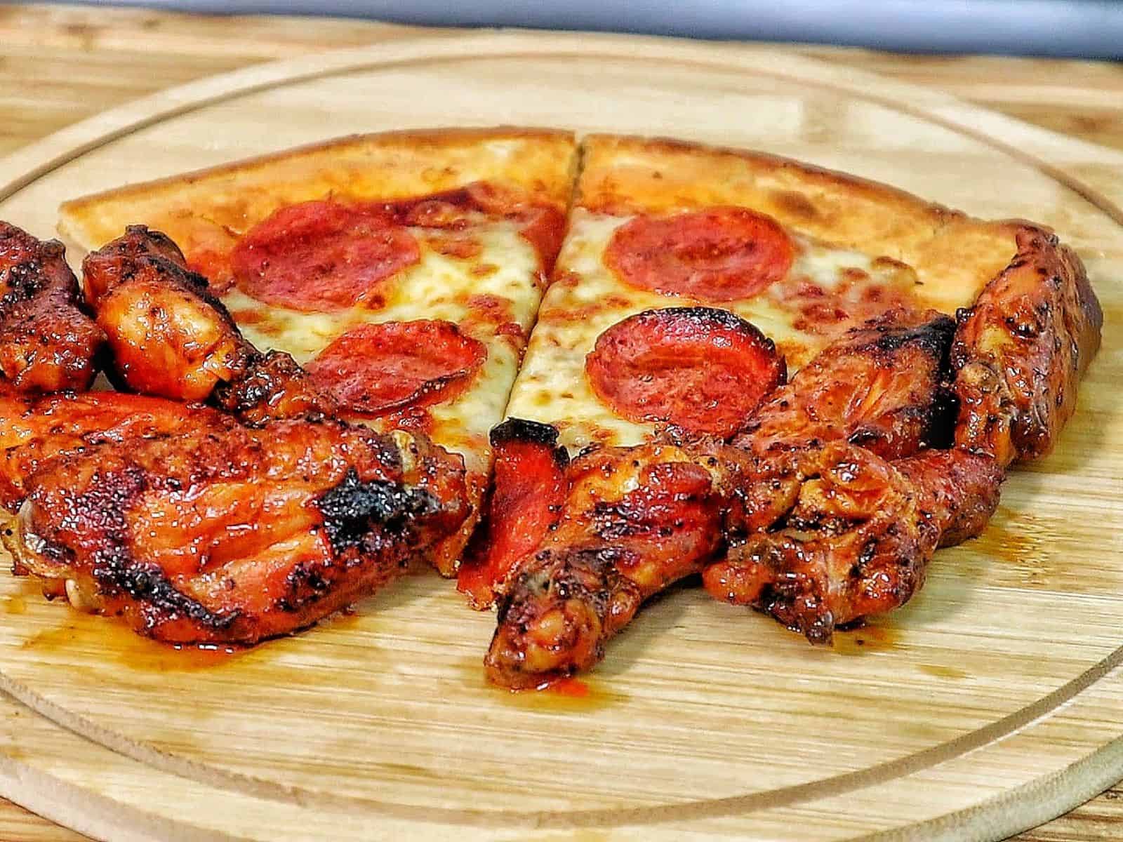 Homemade pepperoni pizza with melted mozzarella and crispy buffalo wings cooked in a pellet pizza oven, served hot with charred crust and golden wings.