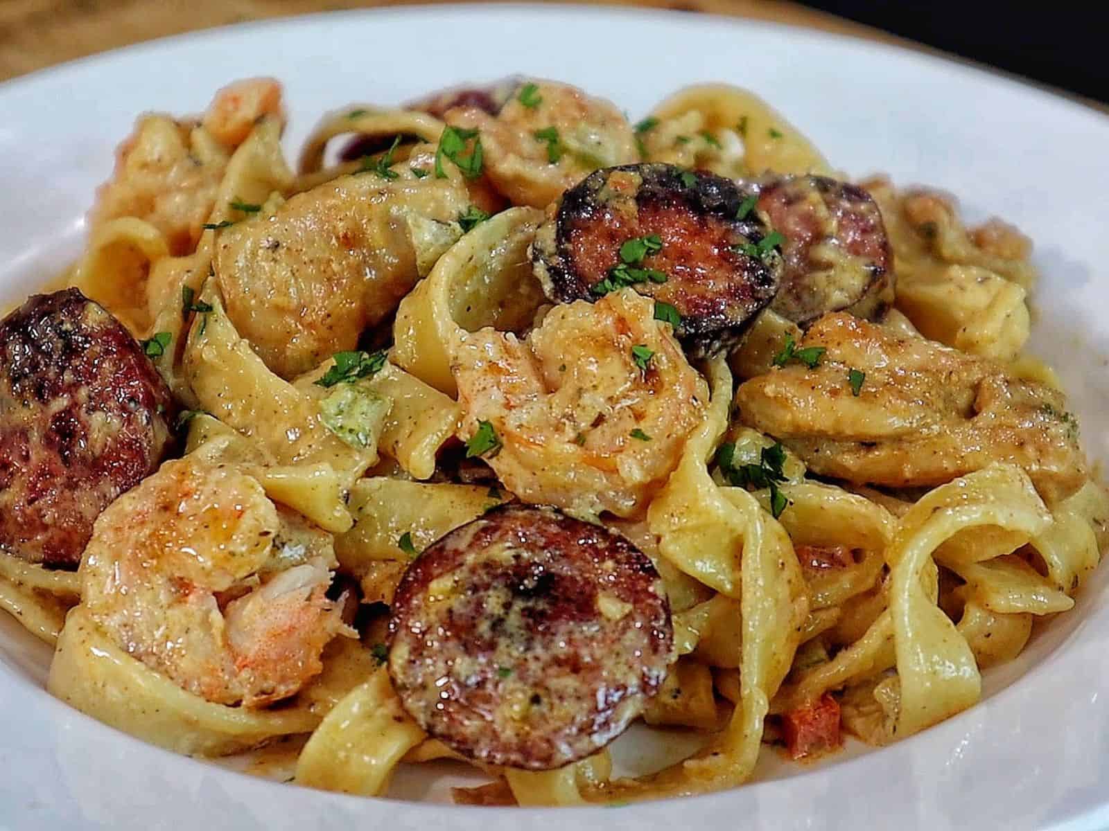 Creamy jambalaya pasta loaded with Cajun-seasoned shrimp, tender chicken thighs, smoked sausage, fettuccine noodles, peppers, celery, onions, and Parmesan cream sauce, garnished with fresh parsley.