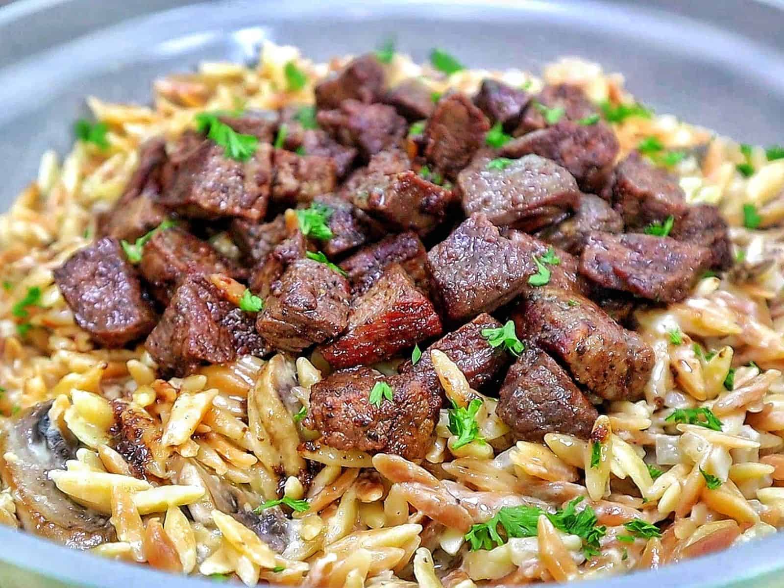 A bowl of creamy Parmesan mushroom orzo topped with perfectly seared steak bites and garnished with fresh parsley