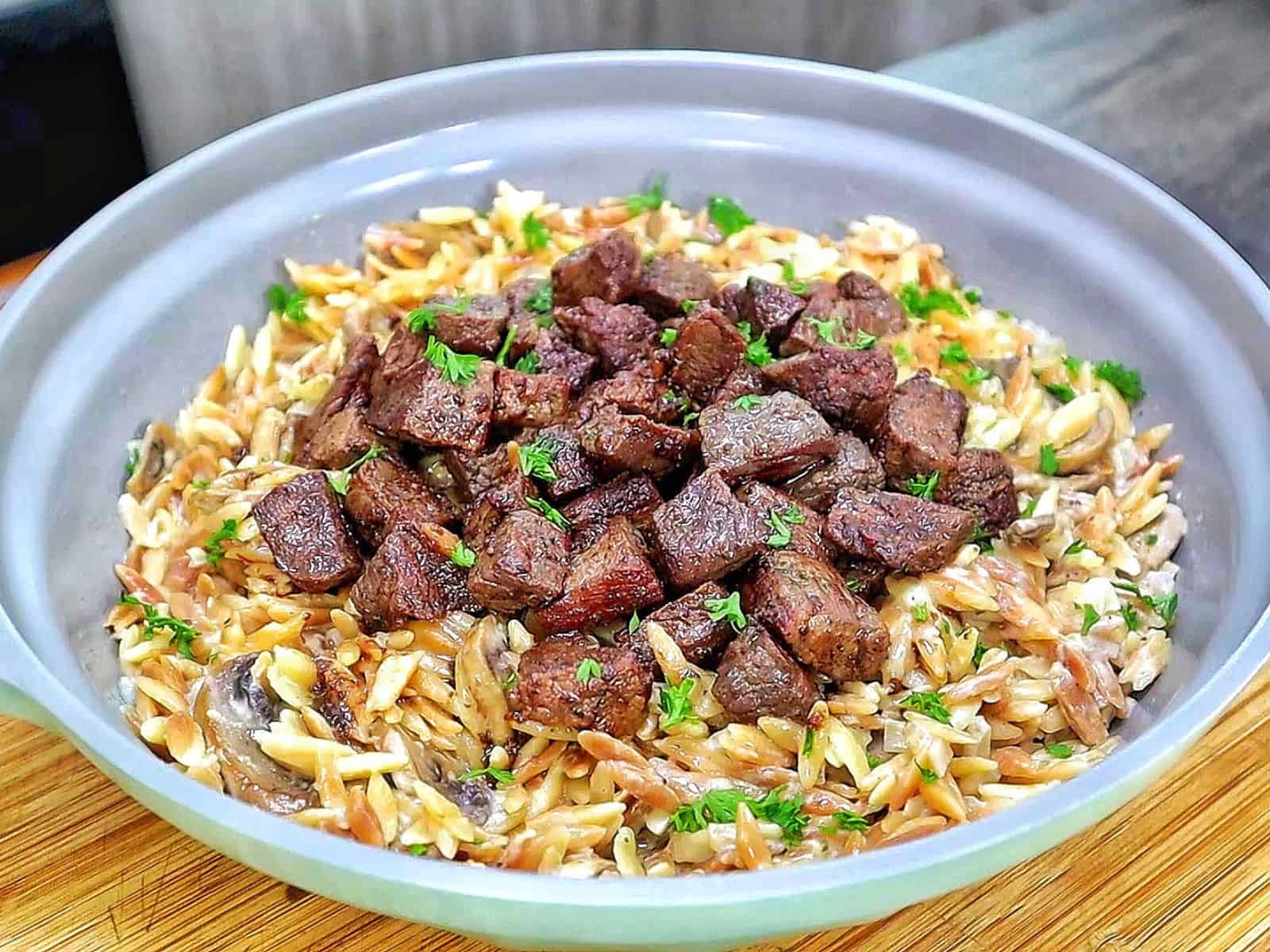 A bowl of creamy Parmesan mushroom orzo topped with perfectly seared steak bites and garnished with fresh parsley