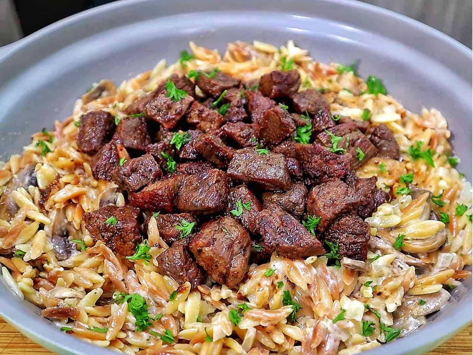 A bowl of creamy Parmesan mushroom orzo topped with perfectly seared steak bites and garnished with fresh parsley