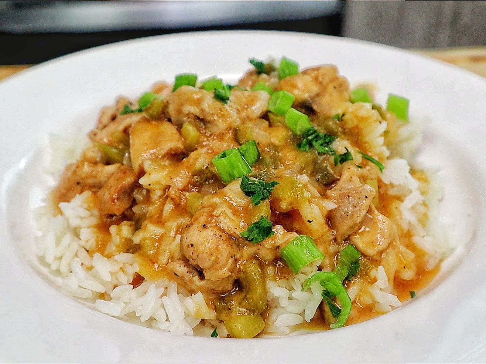 A bowl of homemade Chicken Étouffée served over fluffy white rice, with tender Cajun-seasoned chicken in a rich brown gravy with onions, celery, bell peppers, and topped with fresh green onions and parsley.