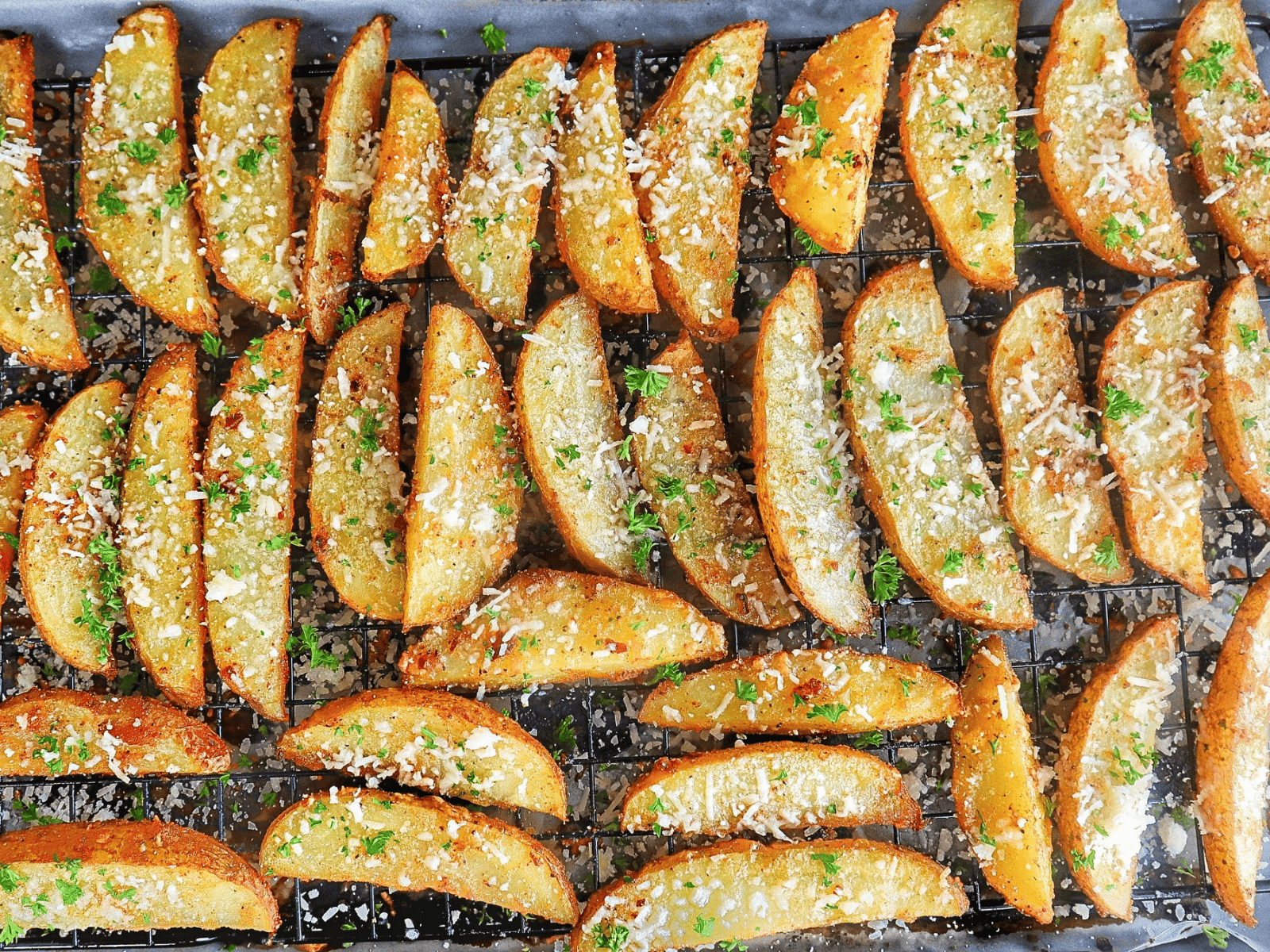 Golden-brown Garlic Parmesan Potato Wedges topped with freshly grated Parmesan cheese and chopped parsley, arranged on a wire rack over a baking sheet.