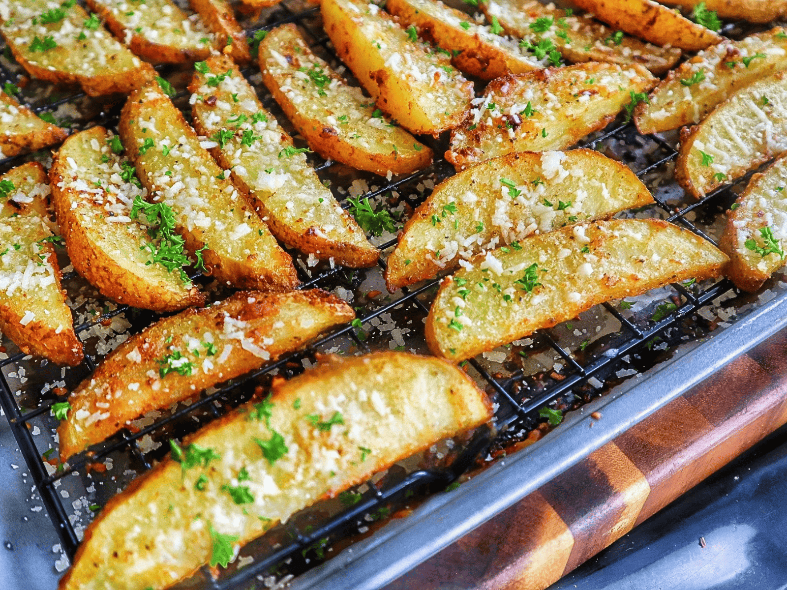 Golden-brown Garlic Parmesan Potato Wedges topped with freshly grated Parmesan cheese and chopped parsley, arranged on a wire rack over a baking sheet.