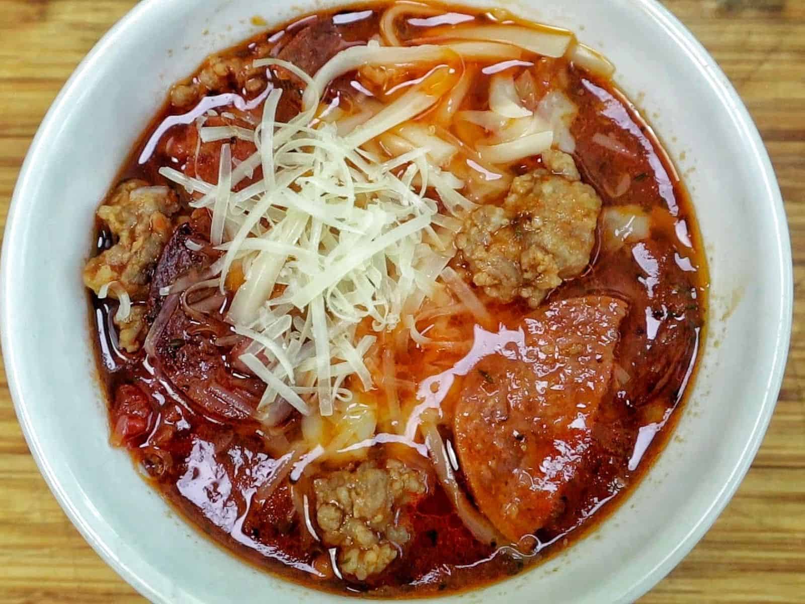 Homemade Meat Lovers Pizza Soup in a white bowl, loaded with Italian sausage, pepperoni, bacon, rich tomato broth, and shredded mozzarella and Parmesan cheese.