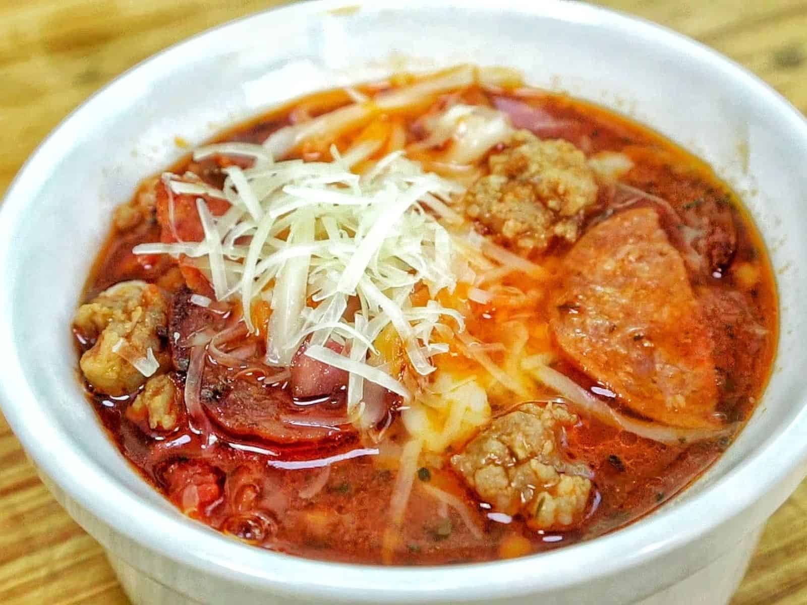 Homemade Meat Lovers Pizza Soup in a white bowl, loaded with Italian sausage, pepperoni, bacon, rich tomato broth, and shredded mozzarella and Parmesan cheese.