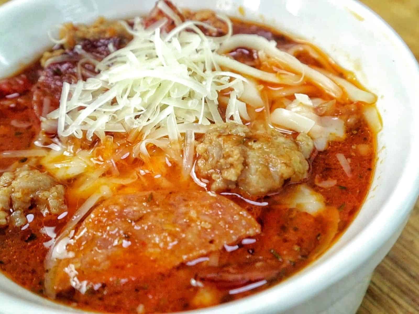 Homemade Meat Lovers Pizza Soup in a white bowl, loaded with Italian sausage, pepperoni, bacon, rich tomato broth, and shredded mozzarella and Parmesan cheese.