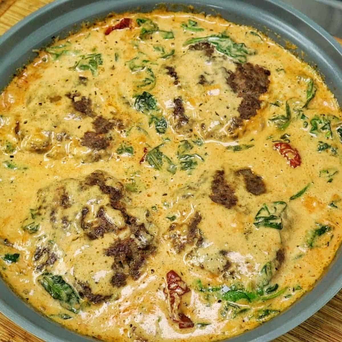 Pan-seared Tuscan Hamburger Steaks simmering in a rich Parmesan cream sauce with spinach, garlic and sun-dried tomatoes in a large skillet.